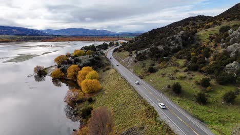 a drone smoothly flies over a winding lakeside road, following a car past autumn trees and rocky hills under overcast skies, with water reflections visible