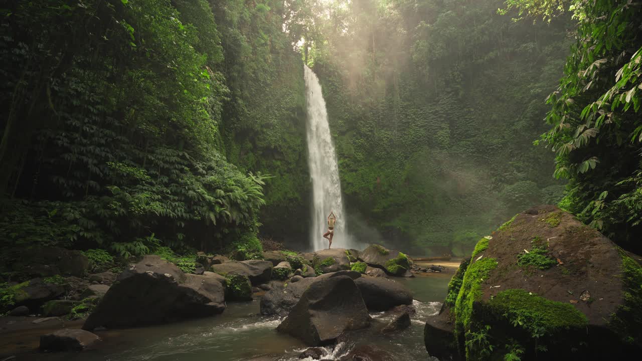 mujer practicando pose de árbol en roca en la magnífica cascada nungnung con luz solar mágica