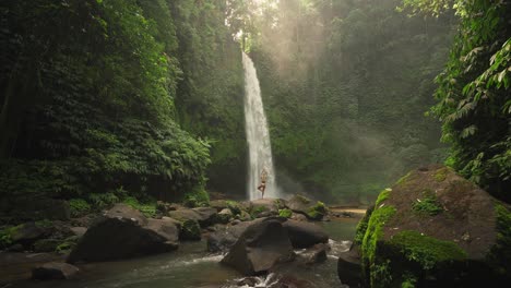 Mujer-Practicando-Pose-De-árbol-En-Roca-En-La-Magnífica-Cascada-Nungnung-Con-Luz-Solar-Mágica