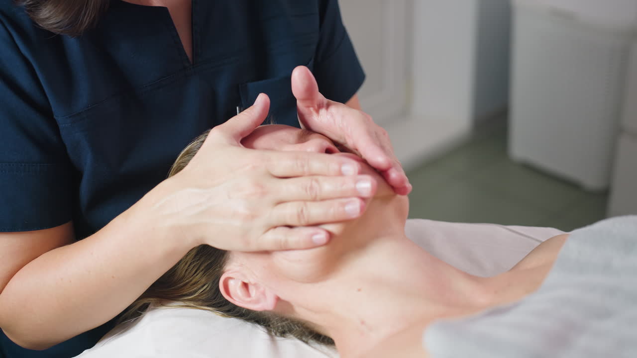 Technician massages forehead of relaxed client lying on bed under soft lighting with gentle strokes for skin rejuvenation relaxation and wellness during professional facial session