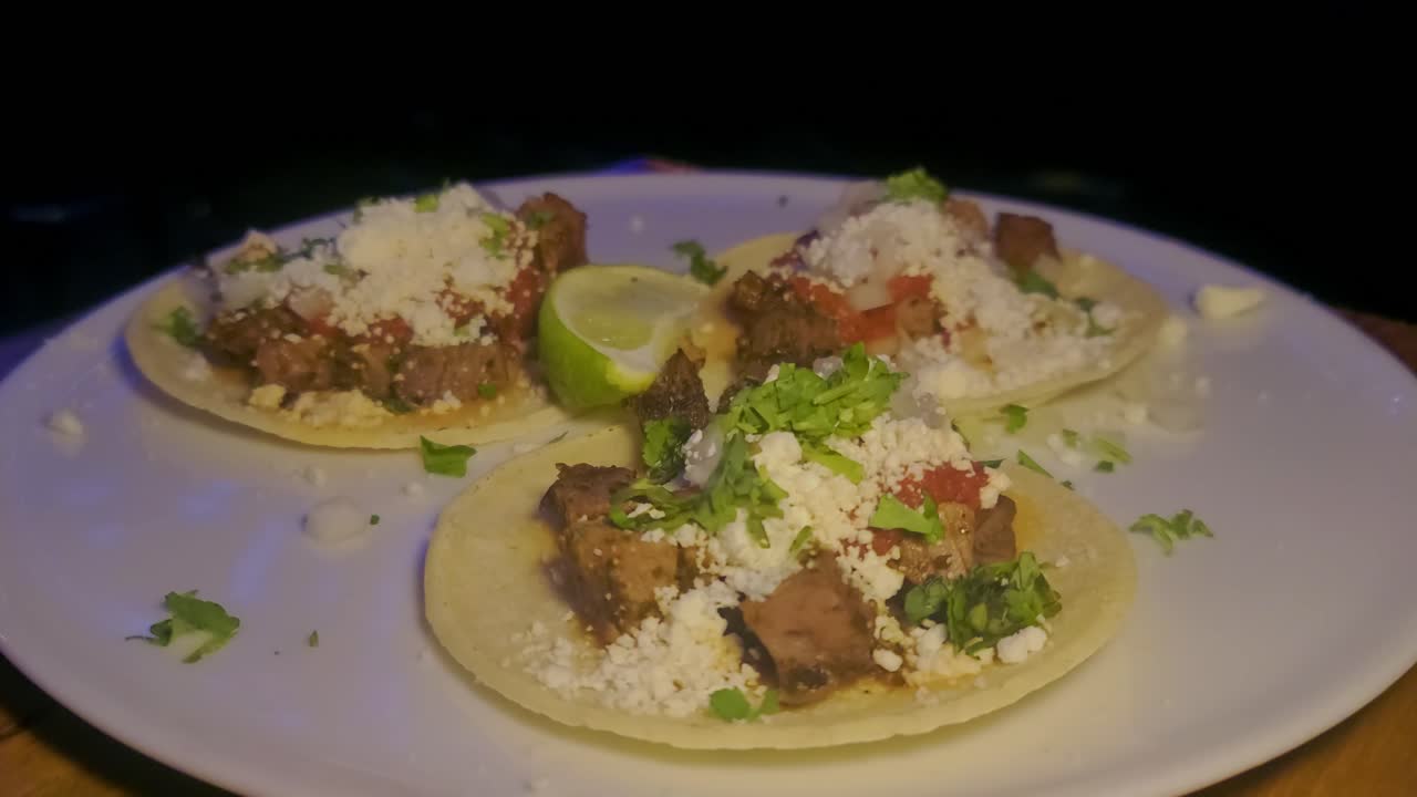 3 tacos de carne zoom óptico de primer plano tortilla de maíz desplegada con cebollas verdes, perejil, limón, salsa en un plato blanco en la parte superior de la mesa de madera con movimiento de fondo oscuro