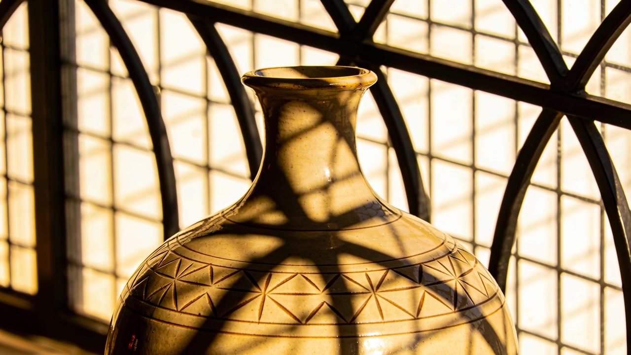 Shifting sunlight casting arched grille shadows across clay vase at sill, showing triangular band