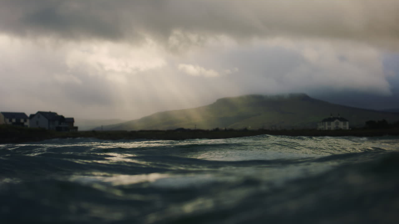 In water slow motion POV of ocean and distant hills as sun light beam rays breaks through dense storm clouds over calm village
