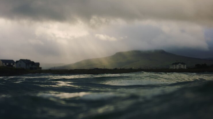 In water slow motion POV of ocean and distant hills as sun light beam rays breaks through dense storm clouds over calm village