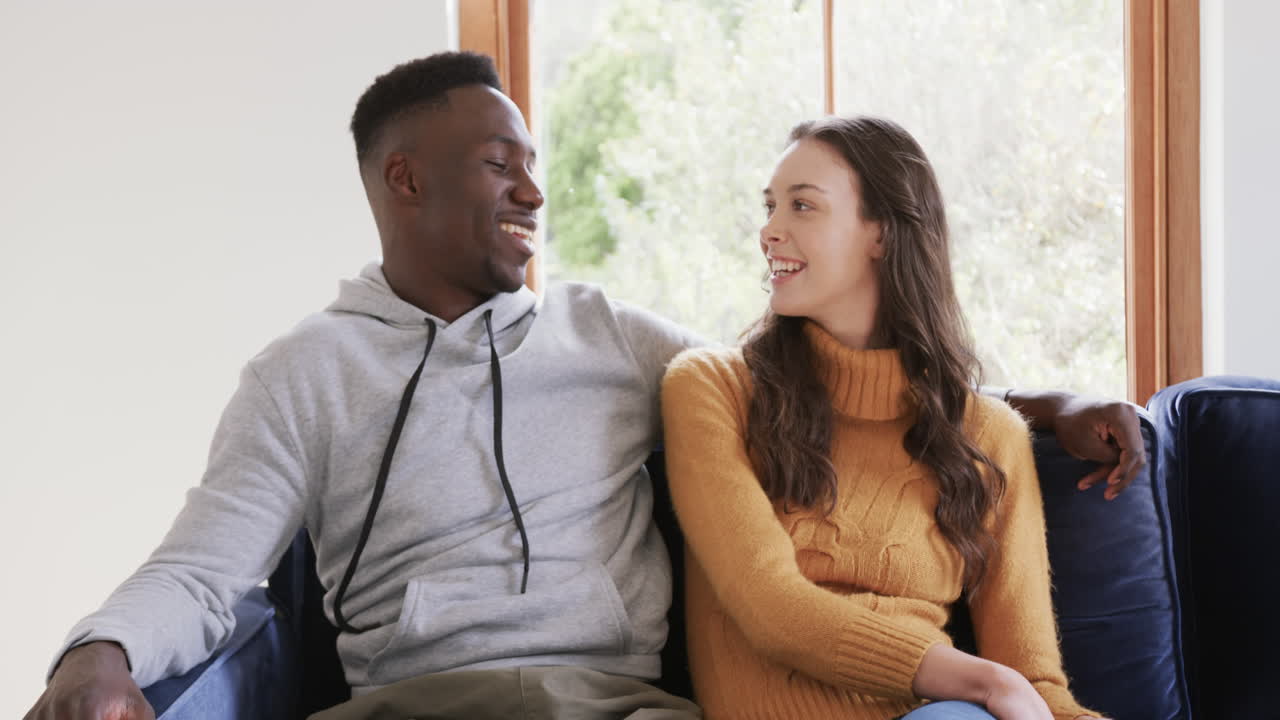 Happy diverse couple sitting on sofa talking and smiling in home,copy space