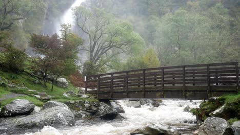 Idyllische-Holzbrücke-über-Dem-Malerischen-Landschaftstal-Wasserfall,-Der-In-Einen-Mächtigen-Fließenden-Fluss-Kaskadiert