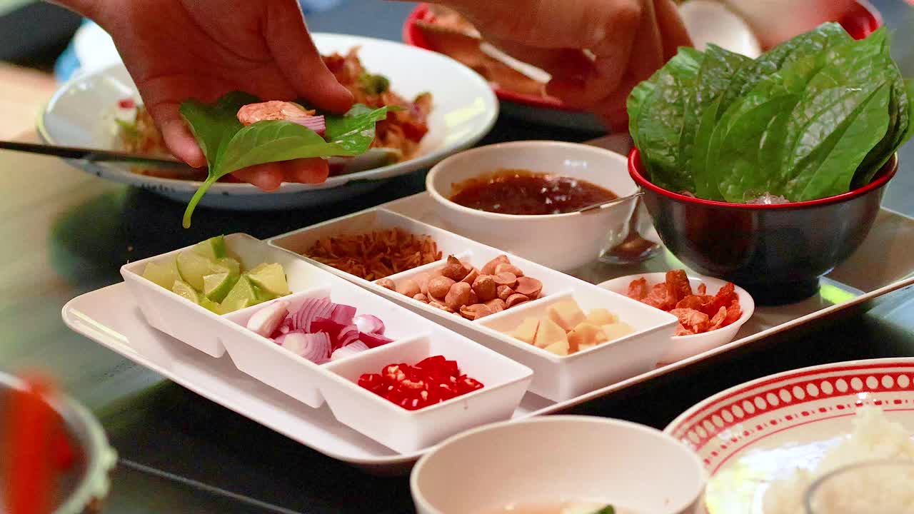 Hands assemble Miang Kham with fresh ingredients on a table in Phuket, Thailand. Bright lighting highlights vibrant colors and textures