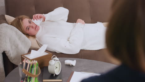 mindful guest lying on sofa during therapy session wiping tears with tissue while speaking to professional, used tissue on table next to pen pack, clock, and notebook in warm counseling room