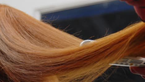 a woman combing a woman's red hair back and then straightening red hair with a straightening iron in a hair salon