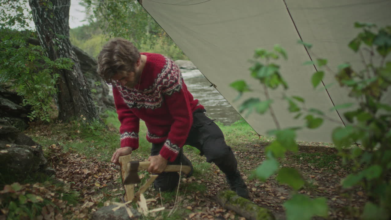 Survivalist Chopping Wood with Hatchet near Tarp Shelter in Forest