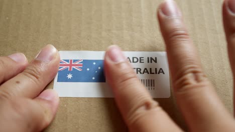 Hände-Bringen-Das-Flaggenetikett-„Made-In-Australia“-Auf-Einem-Versandkarton-Mit-Einem-Barcode-In-Premiumqualität-An