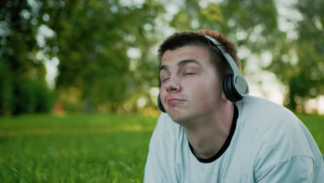 close up of artist lying on grass wearing headphones nodding to music while using phone, gazing up with a subtle smile, lowering his head slightly, surrounded by soft bokeh effect and greenery