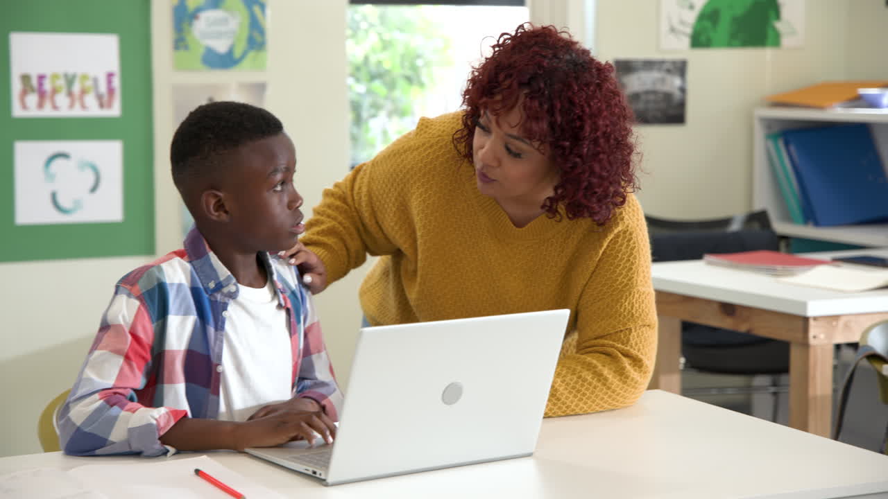 Female teacher helping african american boy with laptop in classroom, focusing on learning, at schoo