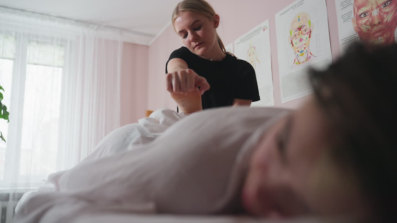 Massage therapist gently lifting client foot toward buttocks during treatment session in tranquil massage clinic, with anatomical charts on wall and blurred view of relaxed client face in foreground