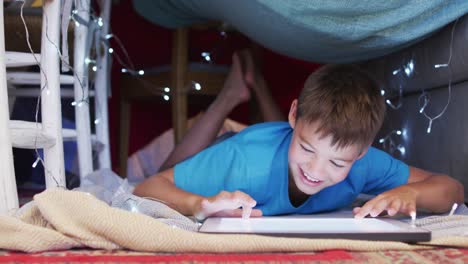 caucasian boy smiling while using digital tablet under the blanket fort at home