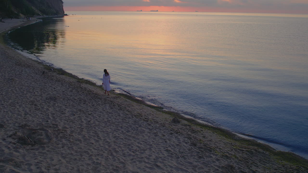 la antena sigue a la mujer caminando en la playa sola mientras el resplandor del atardecer se refleja en el agua