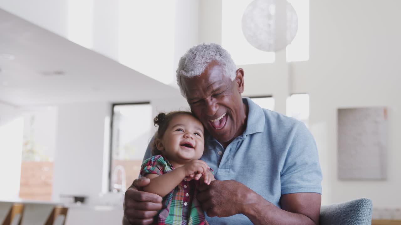 Portrait Of Grandfather Sitting On Sofa At Home Playing Game With Baby Granddaughter