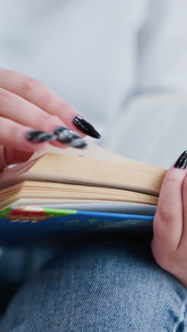 close-up of woman with black nails flipping through pages of an open book while seated with crossed legs, soft bokeh lights create a warm ambiance in the background