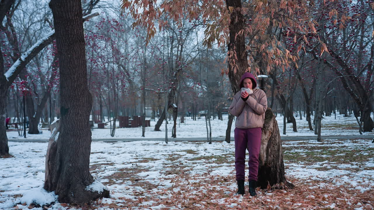 겨울 동안 야외 공원에서 무언가를 먹고있는 나무에 대한 휴식을 취하는 여성, 눈 인 땅, 겨울 재과 후디를 입고, 식사를 즐기고, 겨울 공원 설정, 추운 날씨 휴식