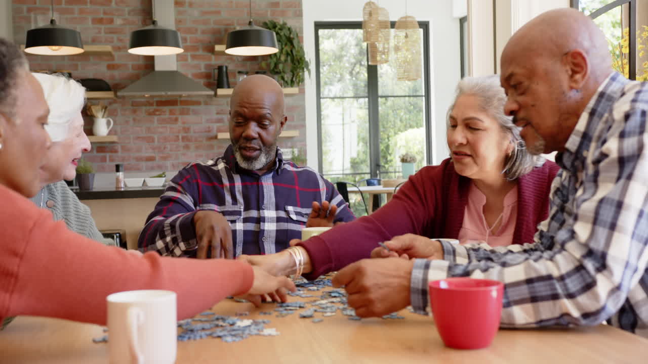 Happy diverse senior female and male friends talking and doing jigsaw puzzle in kitchen, slow motion