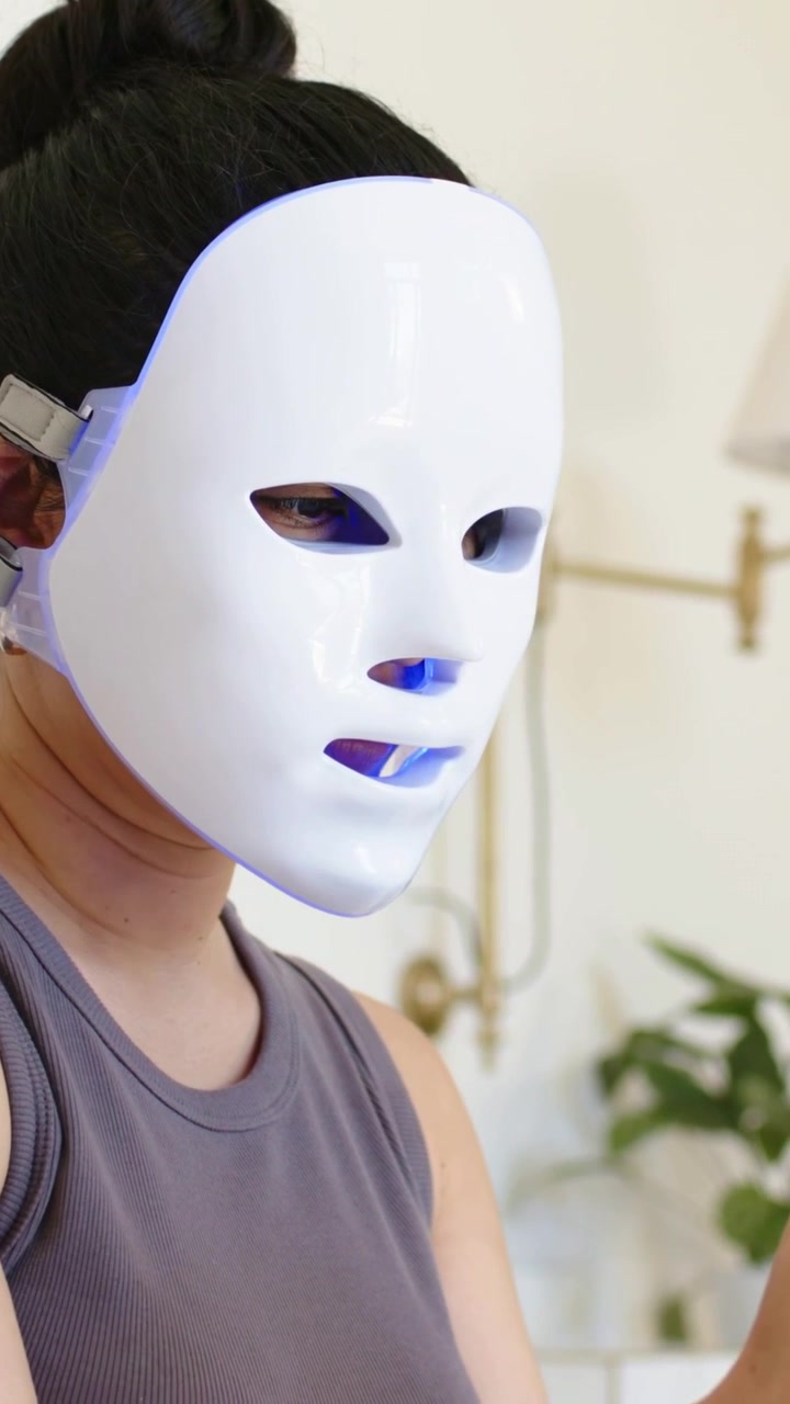 Vertikal video: Kameraet skifter og afslører en kvinde, der tapper på telefonen og justerer en LED-maske, iført grå tanktop derhjemme.