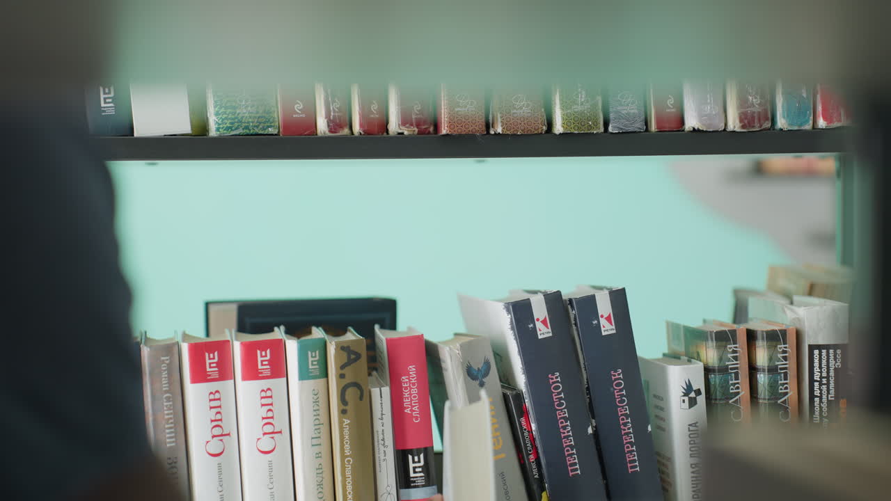 Primer plano de una mano que retira cuidadosamente un libro de la estantería de la biblioteca; los dedos se deslizan por el lomo entre títulos coloridos, ambiente de estudio tranquilo, fondo verde azulado, luz suave, enfoque selectivo, búsqueda de conocimiento capturada en interiores