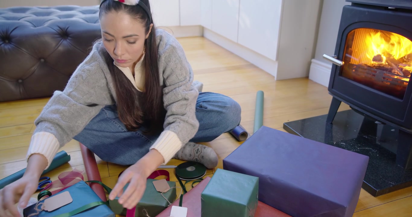 Donna con maglione grigio che prepara regali, iniziando con scatole impacchettate, tagliando il nastro, annodando fiocchi, accanto a una stufa accesa.