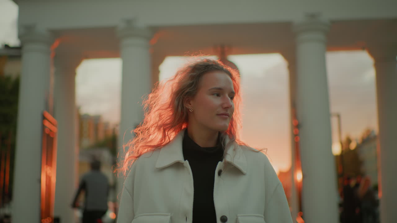 young woman with curly hair wearing white coat looking sideways admiring something in urban environment during beautiful sunset with red glow