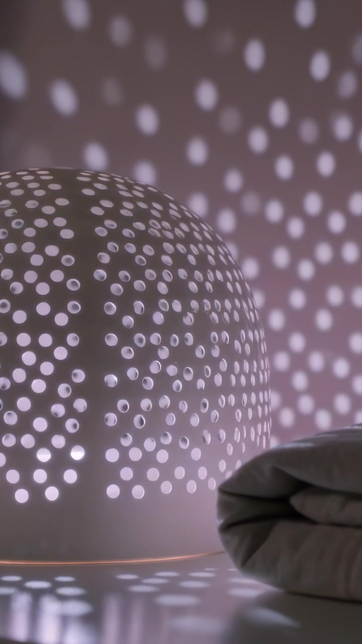 Vidéo verticale : Début d'un cycle de couleurs, lampe perforée projetant des points mouvants sur une étagère.