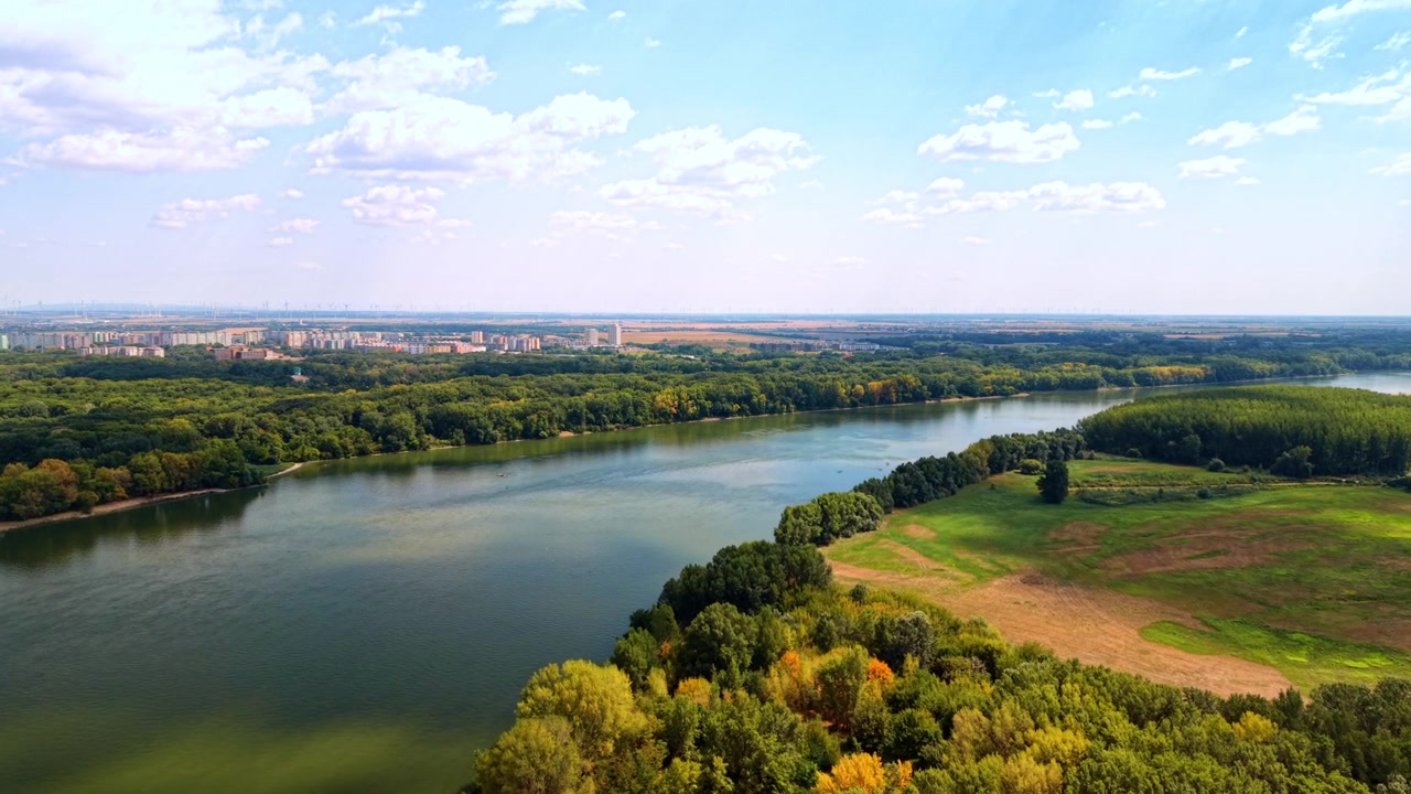 Veduta aerea di un fiume tra boschi e campi aperti. Sponde verdi costeggiano un ampio corso d'acqua accanto a prati a mosaico, con edifici in lontananza sotto nuvole sparse