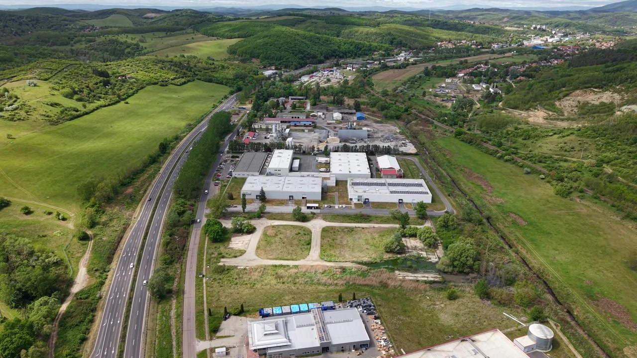 Vista aérea revela edifícios industriais modernos aninhados ao lado de uma rodovia movimentada em uma paisagem verde exuberante e ondulante.