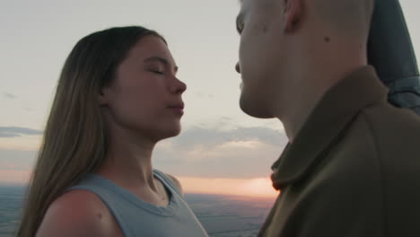 close up of couple sharing intimate gaze while distant red paraglider drifts across pastel sunset sky above sprawling farmland under soft clouds drifting over horizon