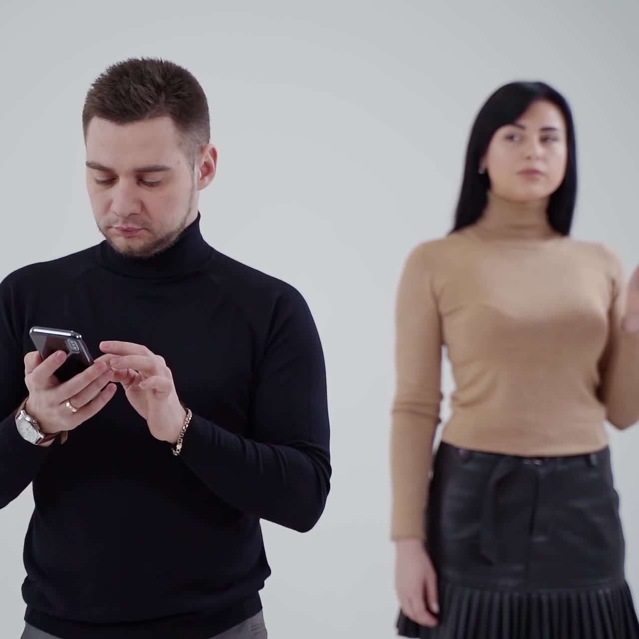 Young man in black sweater clicking the mobile phone. Attractive sad woman standing behind the man and smoking on the studio background.