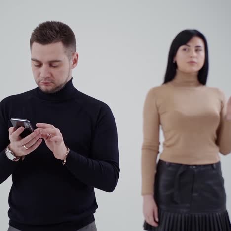 young man in black sweater clicking the mobile phone. attractive sad woman standing behind the man and smoking on the studio background.