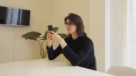 Joven-Con-Anteojos-En-Una-Habitación-Beige-Escribiendo-En-Su-Celular,-Sentado-En-Una-Mesa