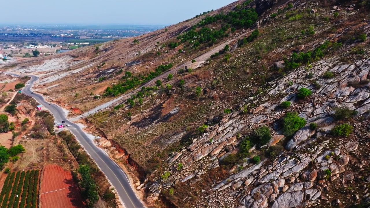 Strada stretta con montagna, terreni agricoli e piante da piantagione sulla strada Isha Adiyogi, Avalagurki, Sonennahalli, Chikkaballapura, Karnataka, India. Di giorno, ripresa con drone, 4K.