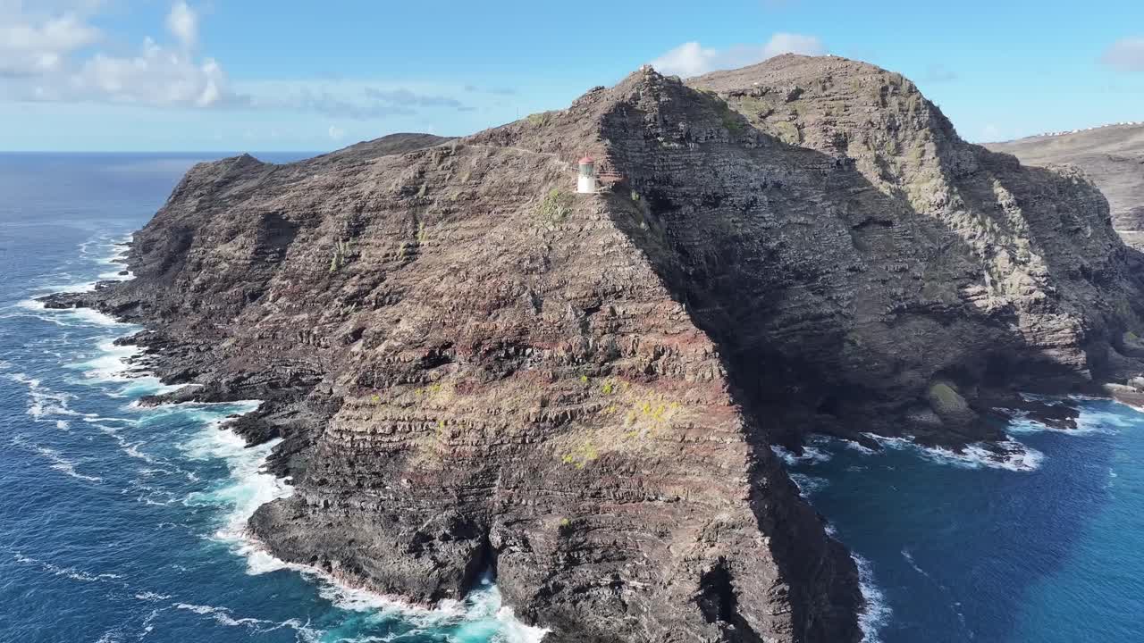 Aerial drone footage of a lighthouse perched on Oahu’s windward coast cliffs, Hawaii, showcasing dramatic rocky coastline, turquoise ocean waves, lush tropical vegetation, and panoramic ocean views