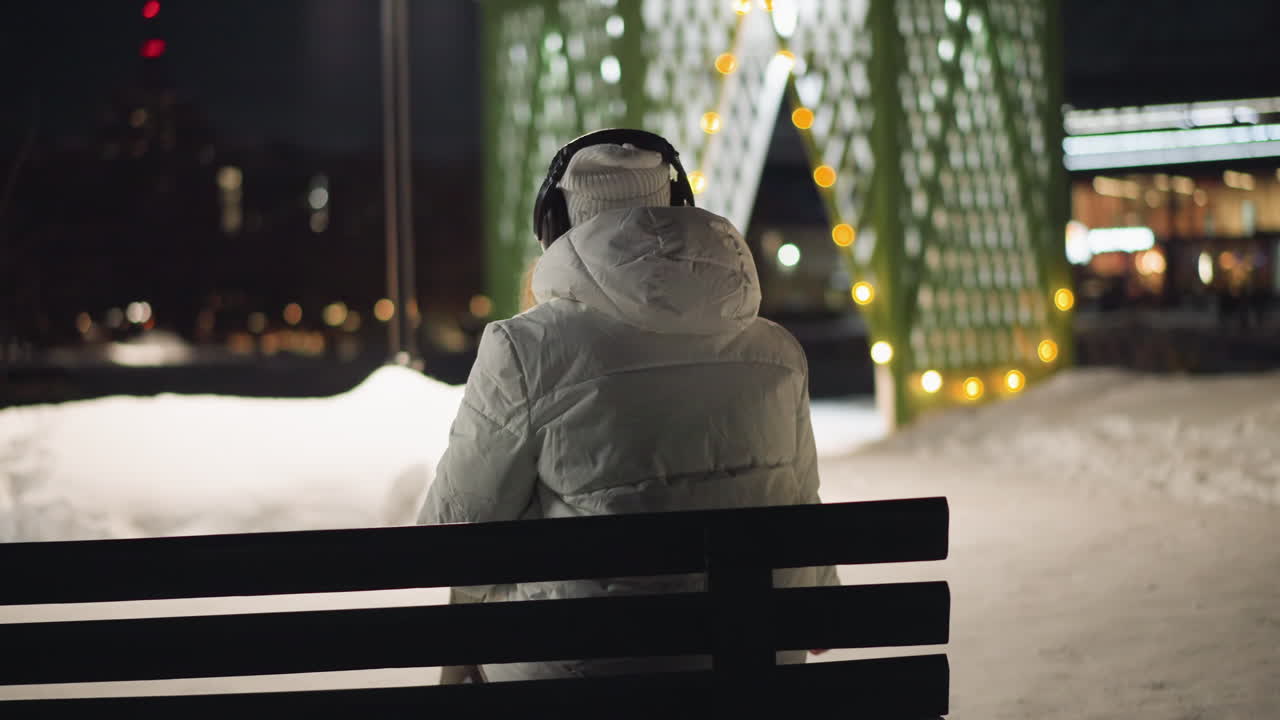 Back view of woman sitting on bench in snowy park at night, shaking head slightly and looking around as festive lights glow behind her, wrapped in winter coat, immersed in calm reflective moment