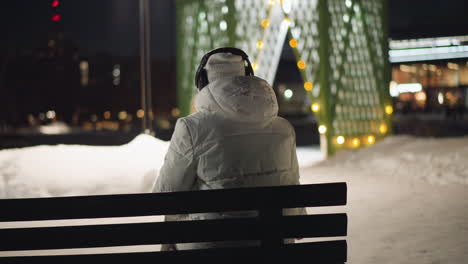 back view of woman sitting on bench in snowy park at night, shaking head slightly and looking around as festive lights glow behind her, wrapped in winter coat, immersed in calm reflective moment
