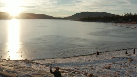 Aerial-flyover-waving-woman-silhouetted-on-rocky-pier-alongside-a-frozen-lake-at-sunset-hour