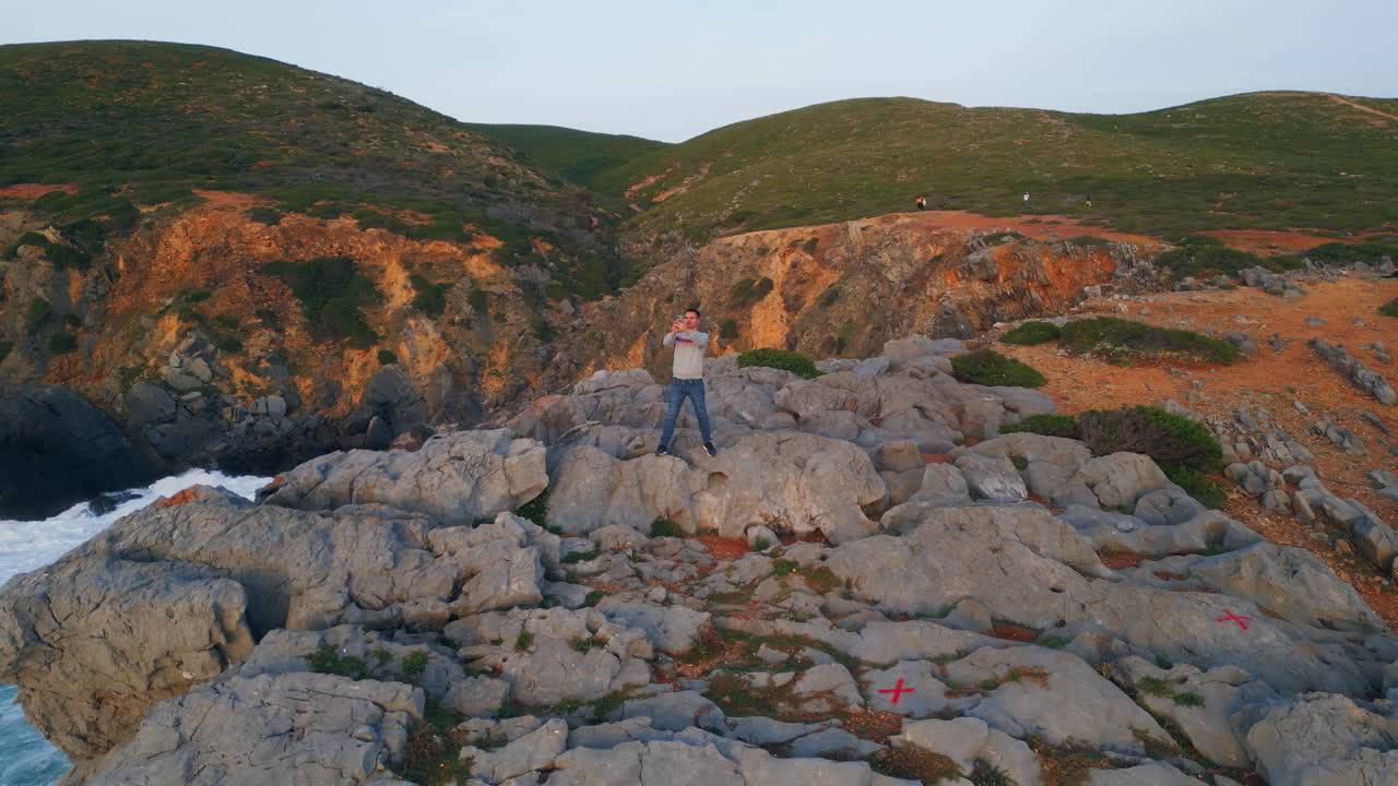 hombre de pie en el acantilado fotografiando la naturaleza de la orilla del mar. colinas verdes entorno marino