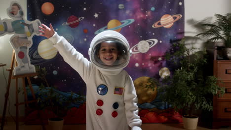 A happy child in an astronaut costume smiles and waves in front of a space-themed backdrop