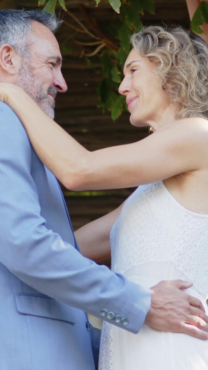 Video verticale: Coppia con le teste vicine che apre gli occhi, chinata sotto un pergolato a un matrimonio, lei in abito bianco.