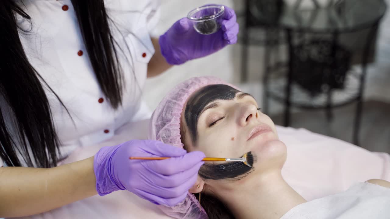 A doctor-cosmetologist applies a black mask with a brush to cleanse and moisturize the skin of the face in the beauty center. Close-up.