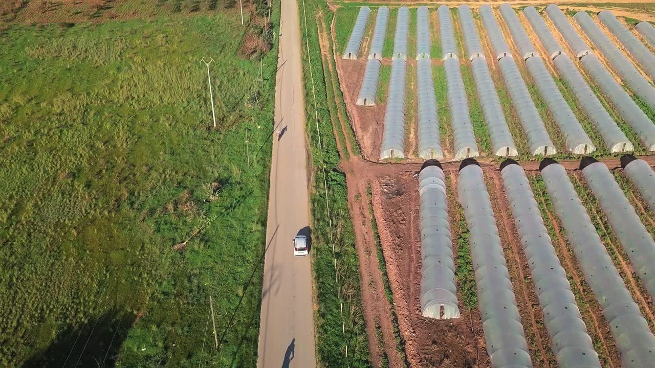 색 자동차가 시골 도로를 지나 들판과 온실을 지나가는 드론 영상