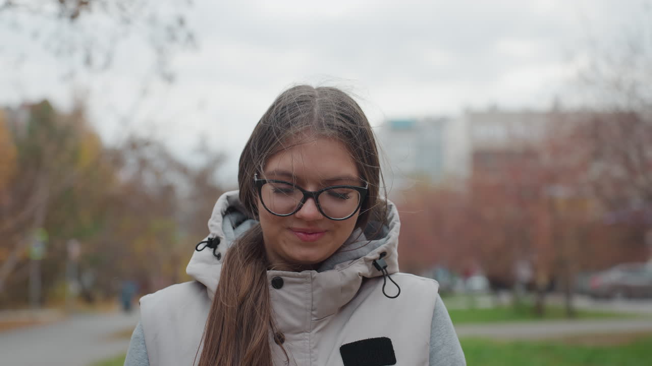 Elegant lady wearing glasses and ash jacket walking with a smile in urban park setting with autumn trees, green grass, moving cars, and residential buildings in background