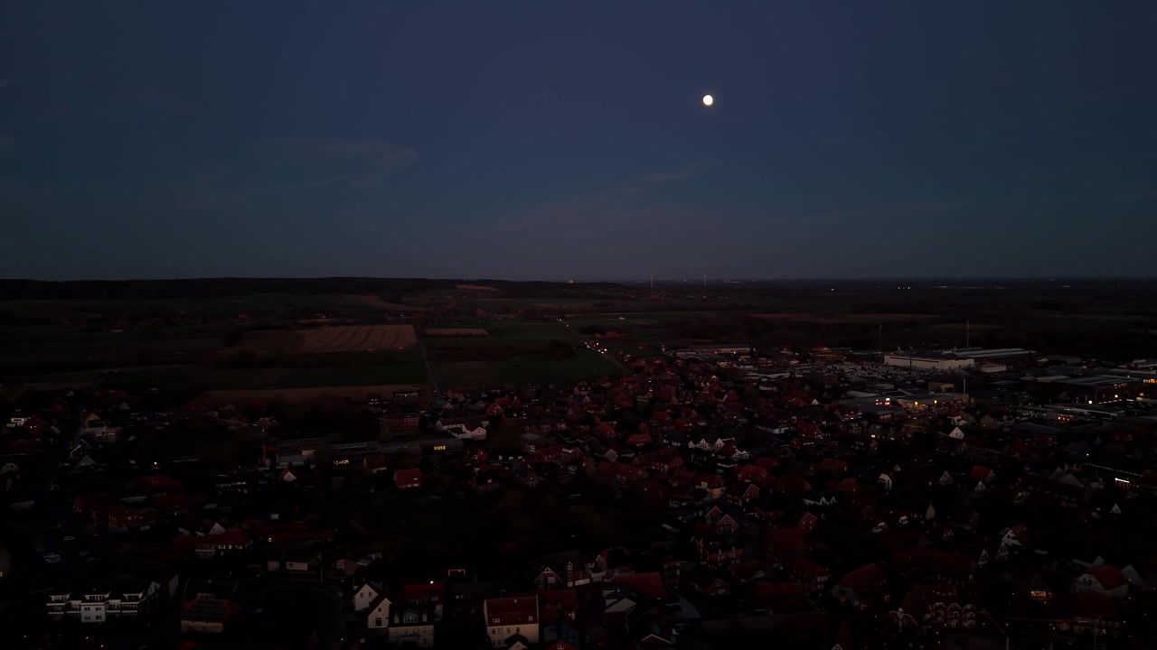 Cittadina illuminata da luci sparse sotto una luna piena luminosa che sorge in un cielo crepuscolare scuro sopra una zona rurale degli Stati Uniti, terreni agricoli distanti si estendono oltre i densi tetti residenziali vicini. Ripresa aerea con drone