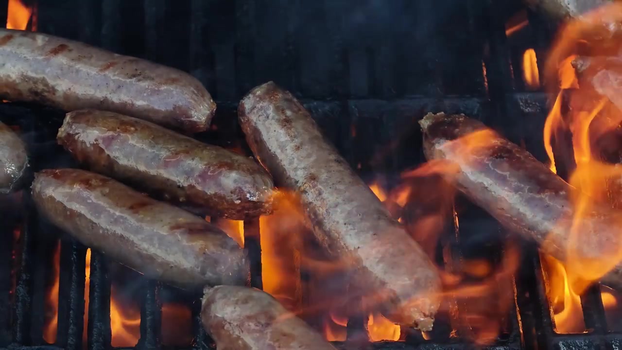 Primo piano in slow motion di salsicce di manzo premium e chorizo piccante che grigliano su fiamme libere su un barbecue nero con fumo drammatico, grasso che sfrigola e scintille di fuoco cinematografiche, riprese dinamiche in 4K del barbecue!