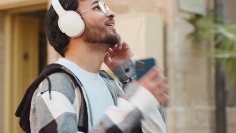 happy indian man wearing headphones choosing listening disco rock n roll music dancing on street