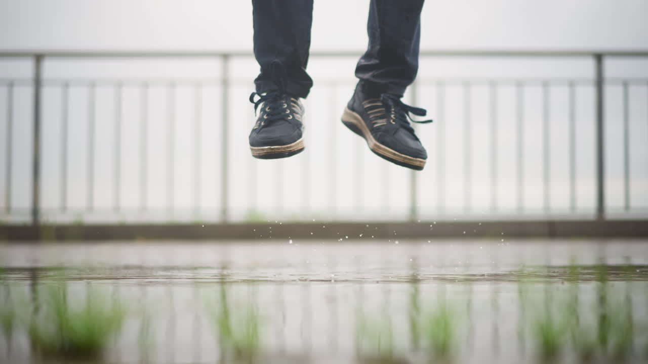 Salto con salpicadura, zapatillas en el aire sobre un charco: un chico blanco juguetón da saltos enérgicos y salpica agua en un paseo por la azotea; gotas de agua y movimiento en el aire; juego de pies de un bailarín callejero que se combina con un estallido atlético.
